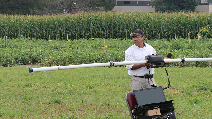 A University of Nebraska–Lincoln researcher stands in a crop field holding a large sensor instrument connected to a laptop, preparing to collect geophysical data to assess soil and water conditions near an irrigation research site.