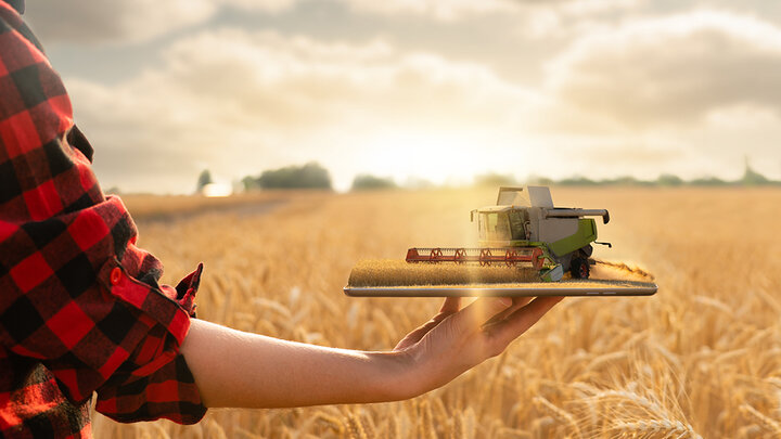 Person in a plaid shirt holds a tablet displaying a three-dimensional combine harvester working in a wheat field under a golden sunset.