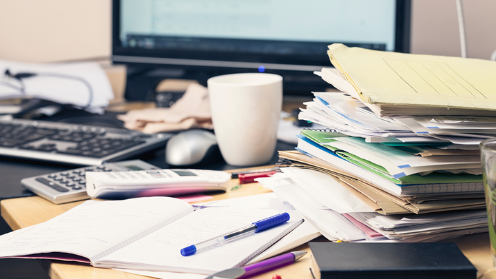 A cluttered office desk with stacks of papers, notebooks, pens and a coffee mug in front of a computer monitor, showing a messy workspace.