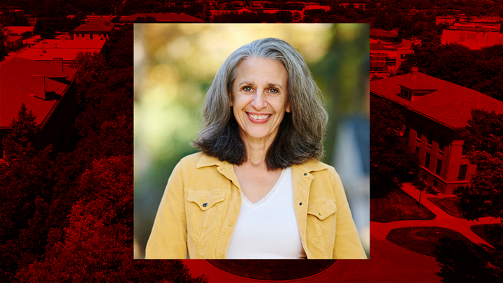 Portrait of a smiling woman with shoulder-length gray hair, wearing a yellow jacket, over a red-tinted aerial campus background.
