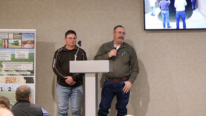 Two men stand at a podium during a conference presentation, one speaking into a handheld microphone, with a slide and dry bean display behind them.