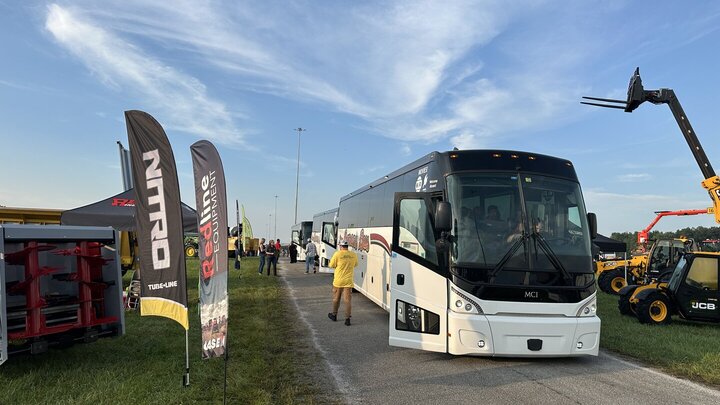 Bus parked on a paved road at an outdoor event with flags and machinery nearby.