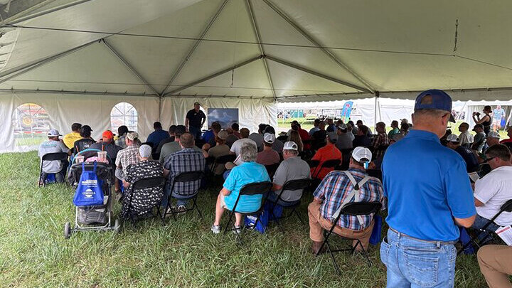 Audience seated under a large white tent on grass, listening to a speaker at the front.