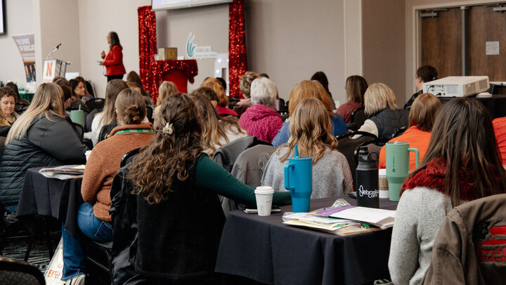 Women attend a Nebraska Women in Agriculture Conference session, seated at tables and listening to a speaker during a large-group presentation.