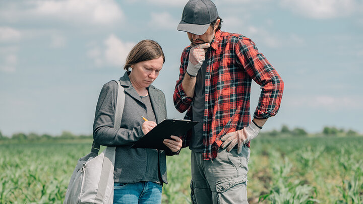 An agricultural specialist and a farmer stand in a green crop field, reviewing notes on a clipboard under a partly cloudy sky. The specialist writes while the farmer, wearing a red plaid shirt and cap, looks on thoughtfully.