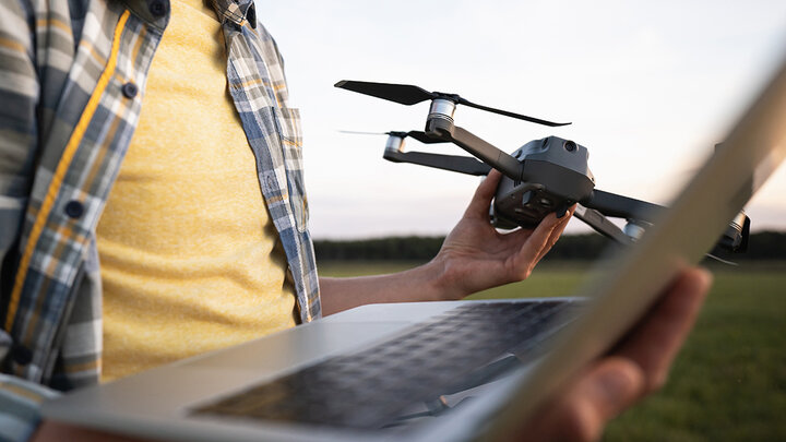 Man holding a laptop in one hand and a small quadcopter drone in the other, standing in an open field, preparing for drone operation or data transfer.
