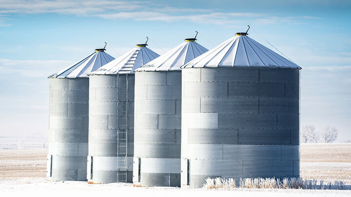 Four silver grain bins stand side by side near a wintry field