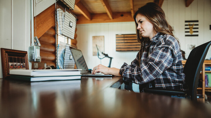 Woman in a plaid shirt works on a laptop at a wooden desk in a home office with shelves and wall art in the background.