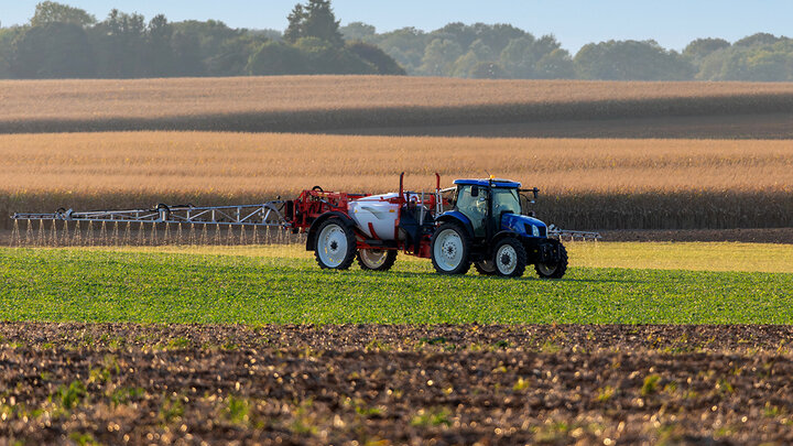 A tractor pulls a boom sprayer across a green field, applying pesticide with mature crops and trees in the background.