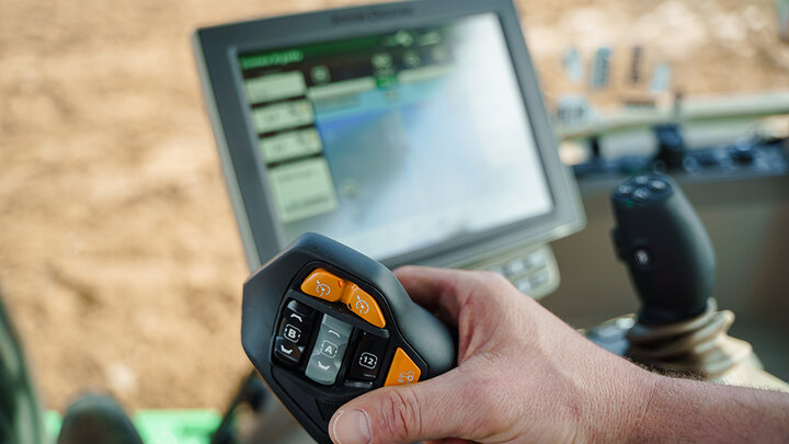 A farmer’s hand holds a precision agriculture control handle inside a tractor cab, with a touchscreen display showing field data and GPS mapping in the background.