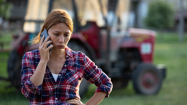 A farmer in a plaid shirt stands outside near a tractor, holding a phone to her ear with a worried expression, reflecting the stress and challenges of farm life.