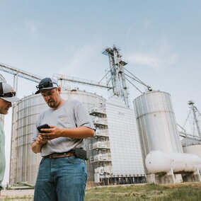 Men look at phone in front of grain silos