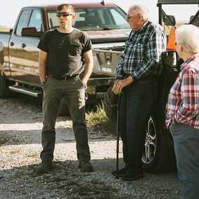 Man speaks to older couple near vehicles and farm equipment