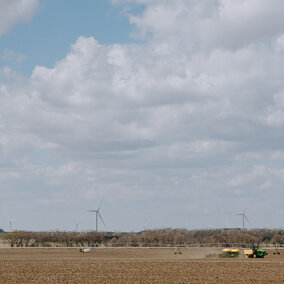 Wide view of Nebraska farmland under a partly cloudy sky, with a tractor and irrigation system working in a freshly tilled field, and wind turbines standing in the distance beyond a line of trees. Ask ChatGPT