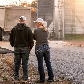 A man and woman in ball caps stand together near grain bins and a semi-truck unloading corn on a farm at sunset.