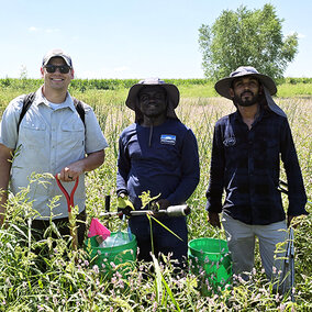 Three men stand in a tall, green grass and wildflower field on a sunny day, holding soil sampling tools and buckets used for soil health benchmarking research.