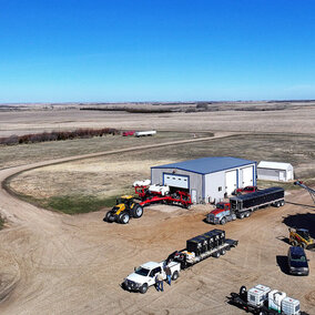Aerial view of a farmstead with grain bins, equipment sheds, semitrucks, tractors, and pickup trucks parked around the yard, surrounded by expansive open fields under a clear blue sky.