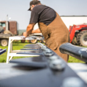 Close-up of a workbench with tools and machine parts laid out in the foreground, while a farmer in brown coveralls performs maintenance on equipment in a field with tractors and harvest machinery in the background