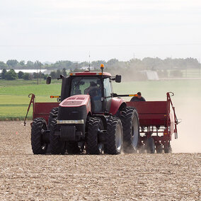 Red tractor pulling farming equipment through a dusty field with green farmland in the background.