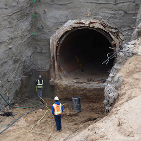 Tunnel excavation site with two workers wearing safety gear, surrounded by dirt and equipment.