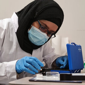 Scientist in lab coat and gloves working with lab equipment at a table.