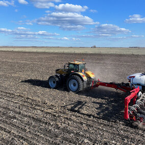 Tractor pulling a red planter across a dry, residue-covered field, kicking up dust under a blue sky with scattered clouds.