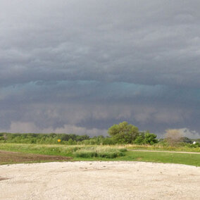 Storm clouds loom over a grassy field with a gravel path and an orange cone in the foreground.
