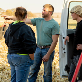 Group talks near truck on harvested field, green equipment in background, sunny day.