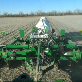 Tractor-mounted applicator injecting anhydrous ammonia into a no-till cornfield, with residue from the previous crop visible on the soil surface and tree line in the background under a clear blue sky.