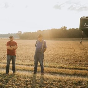 A woman and two men converse in a freshly harvested field near an implement