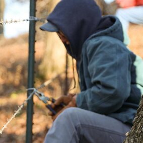 Person repairing barbed wire fence with pliers near a tree in a pasture setting.