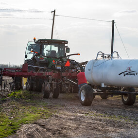 Tractor pulling an anhydrous ammonia tank and applicator toolbar in a field, preparing nitrogen fertilizer for crop production.