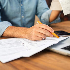 Person reviewing paperwork and receipts with pen at desk beside laptop.