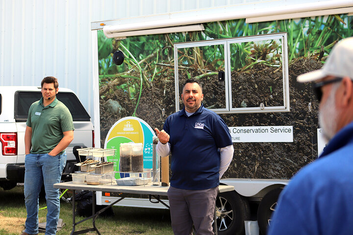 Two men stand beside rainfall simulator while speaking to crowd