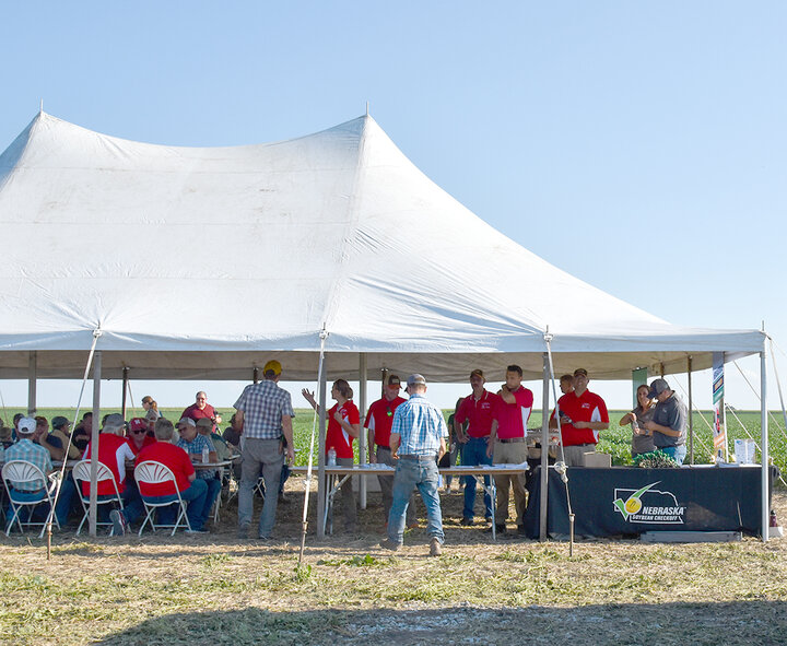Nebraska Extension faculty stand under a large white event tent, speaking with attendees at the 2024 Soybean Management Field Day. Farmers sit at tables inside the tent, and a Nebraska Soybean Checkoff table display is visible in the foreground.