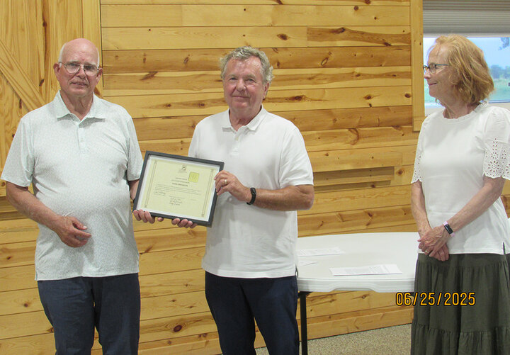 Three people stand indoors as the center recipient holds a framed certificate at an SWCS meeting.