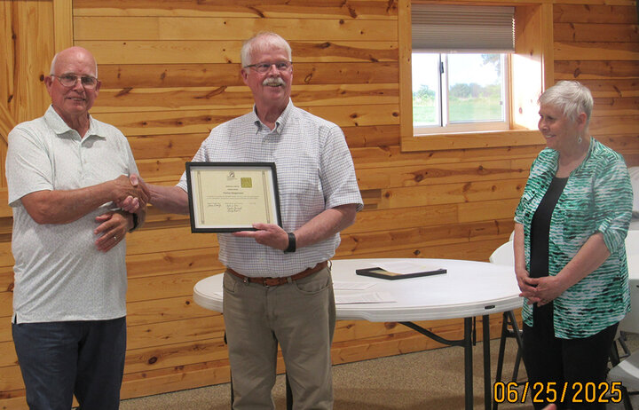 Recipient stands between presenter and guest, holding a framed certificate at a Nebraska SWCS ceremony.