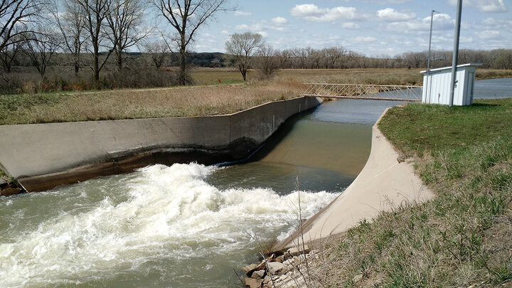 Concrete weir channeling water into a stream with grass and trees in the background.