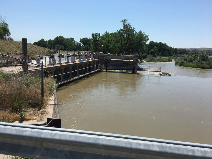 Dam structure with arches spans muddy river, surrounded by trees under a clear blue sky.