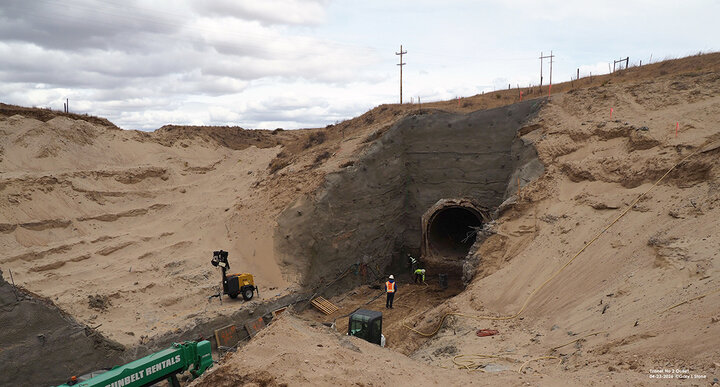 Sandy excavation site with workers near a large tunnel entrance, machinery, and cloudy sky.