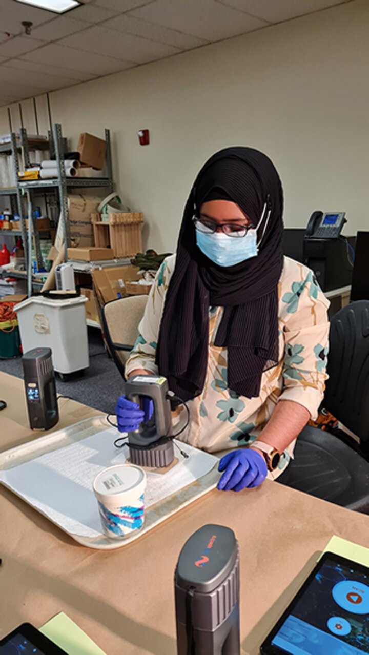 Person wearing gloves using a handheld device in a workspace with trays and equipment.
