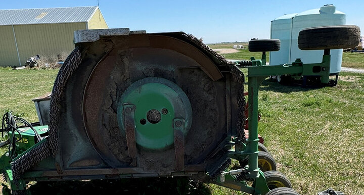 Rusty mower deck in a grassy field with farm buildings and tanks in the background.
