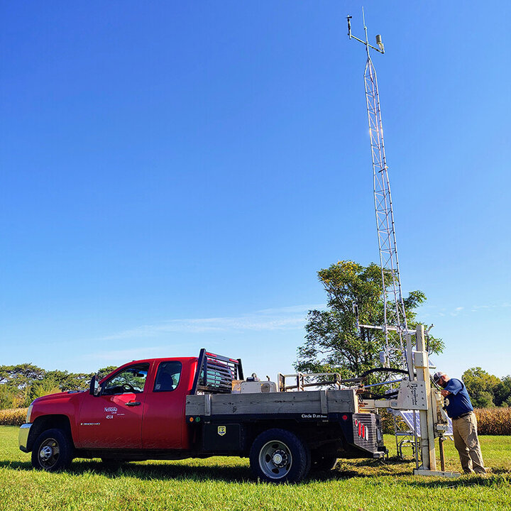 A technician works on a Nebraska Mesonet weather station beside a red university service truck parked in a grassy field under a clear blue sky.