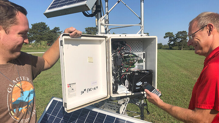 Two men inspect the wiring and equipment inside a Nebraska Mesonet weather station control box in a grassy field.