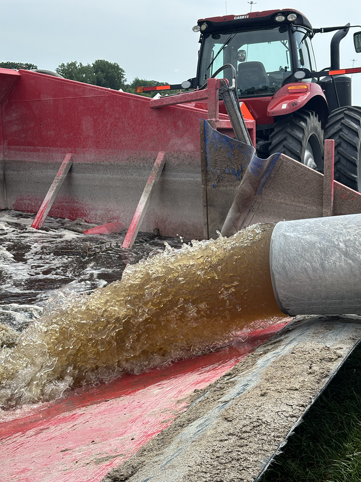 Liquid manure flows from a pipe during a field demonstration, with a tractor and manure handling equipment operating nearby.