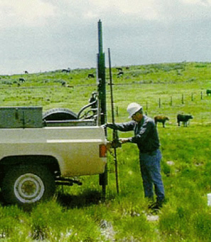 Large pipe apparatus mounted to the end bed of a pickup truck, out in a cow field with man in a hard hat operating.