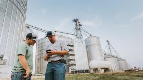 Men look at phone in front of grain silos