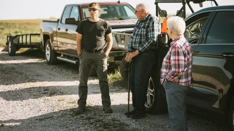 Man speaks to older couple near vehicles and farm equipment