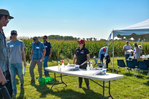 Woman stands at table in front of field day attendees near field